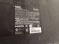 Close-up photo of the NEC model and specification label showing certifications, HDMI logo, Dolby Audio, and serial number.