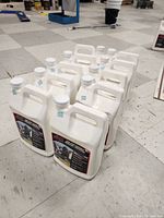 Ten white plastic 1-gallon jugs with handles arranged on floor, showing the quantity and packaging format of the Rejuv asphalt treatment product.
