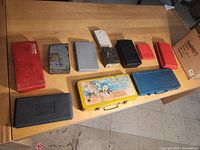 Eleven closed plastic and metal cases of various colors and sizes arranged on a wooden table, mostly rectangular, some labeled.