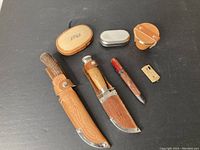 Overall layout showing two knives, two sheaths, wooden box, metal tin, wooden bucket, awl, and brass scraper