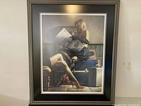 Framed art print showing a woman in a black dress reading a newspaper, sitting on vintage trunks and a chair, captured from a front angle.