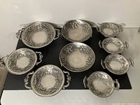 Nine vintage pewter bowls arranged on a black mat, showing varying sizes and consistent grapevine decorative motifs with handles.