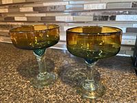 Two entire margarita glasses showing amber-colored bowls, clear stems with bluish green tint at base, set on kitchen counter with tiled backsplash.