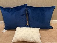Two blue square throw pillows with velvet-like texture and one small white My Pillow travel size pillow on carpeted floor against beige wall.