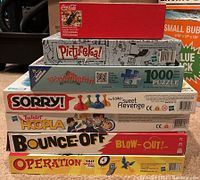 Stacked boxed board games and puzzles arranged for inventory photo including Operation, Bounce Off, Twister Hoopla, Sorry!, Pictureka!, and two 1000-piece puzzles (Washington and Coca-Cola)