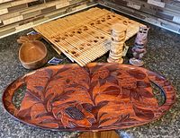 Four bamboo placemats, pineapple monkey pod wood bowl, two tiki statues, and carved wooden tray from Haiti shown together on dark countertop.