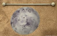 Photo shows a round frosted glass ceiling light cover with purple butterfly designs placed next to a silver adjustable curtain rod with decorative finials on a carpeted floor.