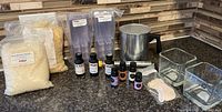 Photo showing candle making supplies arranged on a kitchen countertop including bags of beeswax pellets, tart mold containers, melting pot, temperature gauge, fragrance oil bottles, cotton wick, and glass candle holders.