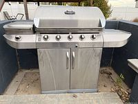 Full front view of the stainless steel Char-Broil 5 burner gas grill showing five control knobs, side stove with cover, and two-door storage cabinet below.
