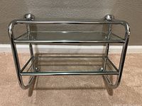 Chrome wall shelf with two glass shelves mounted horizontally, photographed against wall.