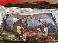 Photo showing Coleman 11x11 screen house being used by a family enjoying a picnic inside the mesh enclosure near natural outdoor setting.