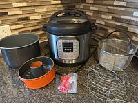 Photo of Instant Pot and all included accessories on a kitchen counter, showing the 6 Qt pot, additional non-stick insert, Bundt pan, two racks, steamer basket, and silicone spout attachments.