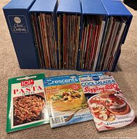 Four blue storage sleeves labeled Pillsbury Classic Cookbooks containing various cookbooks and recipe booklets, with three separate recipe booklets laid out in front.