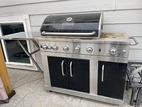 Master Forge gas grill with 5 burners including side sear and rotisserie, two metal side shelves, one foldable, stainless steel control panel with knobs, black front cabinet doors, grill hood with temperature gauge. Grill shows signs of use and one wheel is damaged.