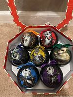 Photo showing all seven Nightmare Before Christmas character-themed Christmas tree ornaments in a decorative box with a transparent lid. Each ornament has a unique character and colored ribbon for hanging.