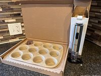 Pampered Chef stoneware muffin pan with 12 muffin cups displayed in open original box on kitchen counter with tile backsplash.