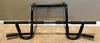 Black metal pull up bar with foam grips resting on floor against wall, showing full structure and handle grips.