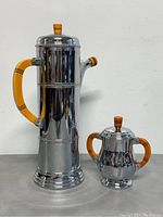 Front view of the chrome coffee carafe and lidded sugar bowl set showing shiny finish and amber Bakelite handles/finials.