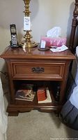 Nightstand with brass lamp, cordless phone, tissue box, and papers on top, showing drawer and open bottom shelf with books.