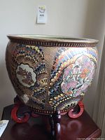 Front view of ceramic planter pot showing painted birds and floral patterns with gold accents and the wooden stand beneath.