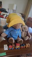 Photo showing a 25 inch Paddington Bear plush, wooden toy train, and plush rabbit on a table.
