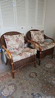 Two matching rattan chairs with floral cushions positioned side by side on a patterned rug in front of white louvered closet doors.