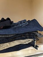 Stack of folded pants showing a mix of denim, corduroy, and lightweight fabric pants in dark blue, black, and gray colors.