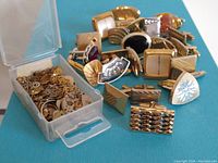 Top-down view of multiple vintage cufflinks spread out next to an open plastic container with assorted watch gears and parts inside.