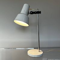 Front angle of white metal desk lamp turned on showing the light emitted and design details of shade and base.