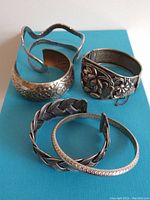 Five distinct vintage silver-tone bracelets arranged on a blue surface showing full lot contents