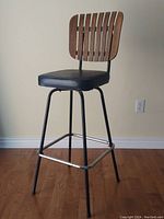 Front view of the mid-century modern swivel bar stool showing wooden slatted backrest, black vinyl seat, metal legs and chrome footrest.