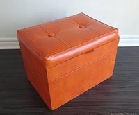 Front angled view of the flame orange vinyl ottoman with button tufting on top and visible vinyl texture.