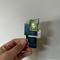 Front view of the MCM stone and silver pinback brooch held in hand showing green and blue rectangular stones set in textured silver-tone metal.