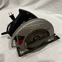 Side view of Skilsaw circular saw on white fabric background showing silver blade guard with logo and black handle with cord.