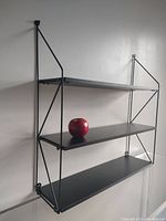 Front view of the black Pilastro style wall shelves mounted on a white wall with an apple on the middle shelf for scale and detail.