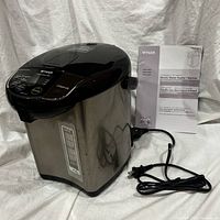 Front angle view of the electric water heater showing stainless steel body, power cord, and manual.