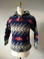 Front view of the woolen ski sweater on a dress form showing the knit pattern with snowflakes and geometric shapes in blue, gray, red, and white, featuring shawl collar and button.