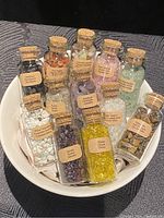 Top view of 12 small glass bottles filled with various crystals placed inside a white ceramic bowl. Each bottle has a label with the crystal name and a cork stopper.