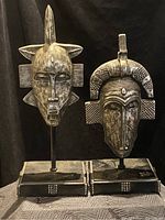 Two carved tribal wooden masks on black stands showing front view, both masks with distinct horn and geometric features.