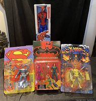 Photo of four action figures in original packaging: Spider-Man boxed, Superman robotic suit, Batman & Robin set, and X-Men figure.