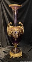 Full view of the large purple glass vase with gold trimmed rim, handles, and base against a black draped background.