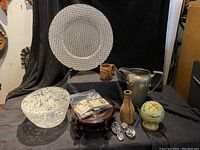 Photo showing the assembled items on a black cloth background including a white decorative plate, pottery items, a metal pitcher, glass bowl, and matchbox