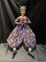 Front view of marionette puppet showing detailed face, ornate headpiece, purple and gold pants, and green feet against black background