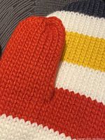 Close-up of the vibrant orange thumb and coloured stripe pattern knit of the mitten.