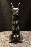 Front view of black indigenous totem sculpture showing detailed facial features and standing pose on base.