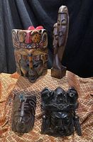 Photo showing all four masks together on patterned fabric background: the colorful mask, the tall twin-faced sculpture, the dark crowned mask, and the horse-themed mask