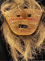 Close-up photo of a woven wicker mask with straw hair and pink accent lines, showcasing facial cutouts