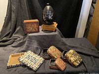 Lot of nine decorative boxes displayed on black fabric, showing the variety including mosaic tile, floral lacquer, metal, and wood boxes.