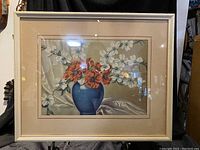 Framed floral painting showing red and white flowers in blue vase on draped background with beige frame.