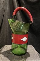 Green blown glass pitcher with red wrapped handle and white diamond shapes on a red band around the middle, shown from front angle.